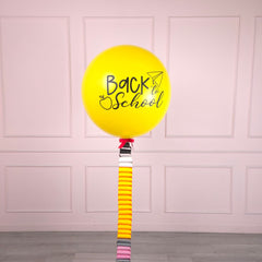 A bright yellow balloon with 'Back to School' printed in black script, attached to a long, striped and yellow pencil-shaped ribbon, set against a light pink paneled wall.