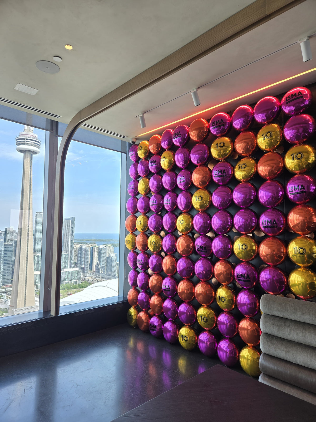 A wall covered in rows of shiny foil balloons in purple, gold, and orange, with a large window showing a city skyline and the Toronto Eye on the left. A gray upholstered chair is partially visible in the foreground.