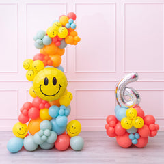A balloon garland featuring a happy face design, with multicolored balloons arranged in a pattern to form a smiley face, standing next to a number 6 balloon, on a pink background.