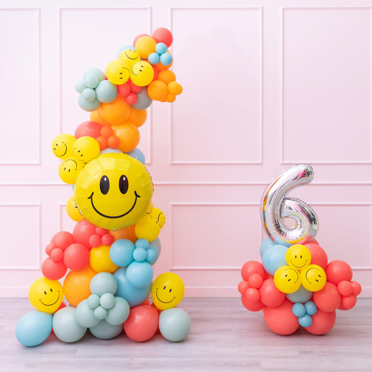 A balloon garland featuring a happy face design, with multicolored balloons arranged in a pattern to form a smiley face, standing next to a number 6 balloon, on a pink background.