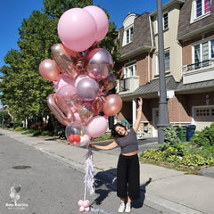 A bunch of pink balloons tied to a balloon pole, with a woman standing on the side of the road holding a balloon.