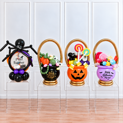 Four Halloween-themed balloon baskets displayed on clear acrylic risers in front of a white paneled wall. The set includes a black spider basket with purple accents, a black pumpkin basket with green and white balloons, an orange jack-o'-lantern basket with multicolor lollipops, and a purple bucket basket with pink and red balloons.