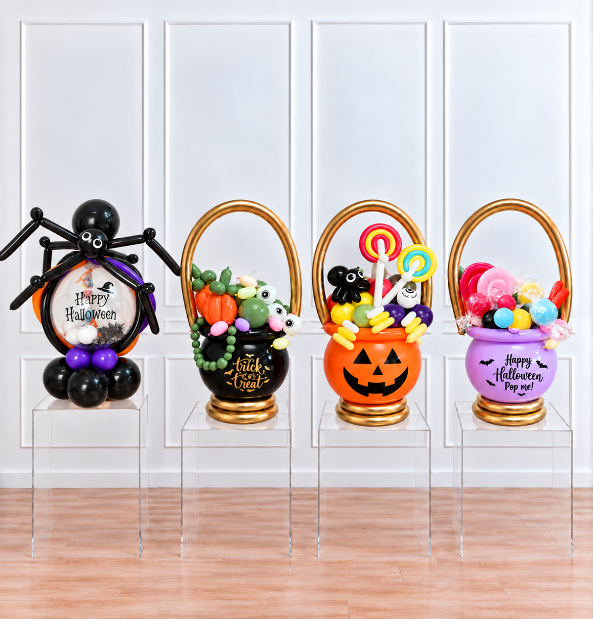 Four Halloween-themed balloon baskets displayed on clear acrylic risers in front of a white paneled wall. The set includes a black spider basket with purple accents, a black pumpkin basket with green and white balloons, an orange jack-o'-lantern basket with multicolor lollipops, and a purple bucket basket with pink and red balloons.
