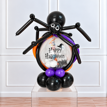 A Halloween balloon sculpture of a spider holding a round balloon filled with 'Happy Halloween' text, black balloons, purple balloons, and small white and black decorations, displayed on a clear acrylic pedestal in front of a white paneled wall.