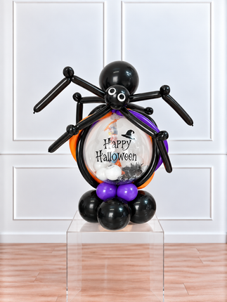 A Halloween balloon sculpture of a spider holding a round balloon filled with 'Happy Halloween' text, black balloons, purple balloons, and small white and black decorations, displayed on a clear acrylic pedestal in front of a white paneled wall.