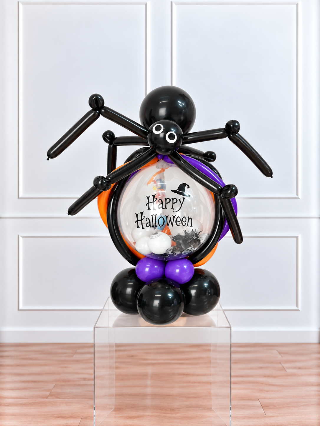 A Halloween balloon sculpture of a spider holding a round balloon filled with 'Happy Halloween' text, black balloons, purple balloons, and small white and black decorations, displayed on a clear acrylic pedestal in front of a white paneled wall.