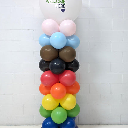 A multicolored balloon column featuring a variety of balloons in different colors stacked vertically, with a prominent pink balloon at the top that reads 'Everyone is Welcome Here'.