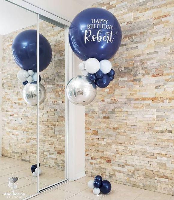 Two large blue balloons with 'Happy Birthday' text and a name 'Robert' printed on them, placed in a room with a brick wall and a mirror.