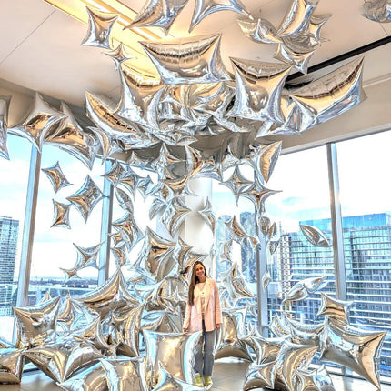 A large number of silver balloons are displayed, forming a decorative arch or structure, with a person standing in front of it.