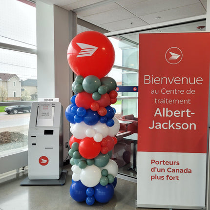 A tall balloon column featuring a variety of round balloons in different colors, including red, blue, green, and white, with a red envelope and the text 'BIENVENUE A CENTRE D'TRAITEMENT A ALBERT-JACKSON' at a kiosk.