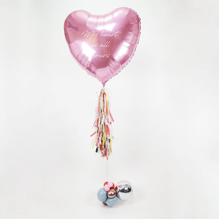 A pink, heart-shaped, jumbo foil balloon with a personalized message, attached to a paper tassel with multicolored elements.
